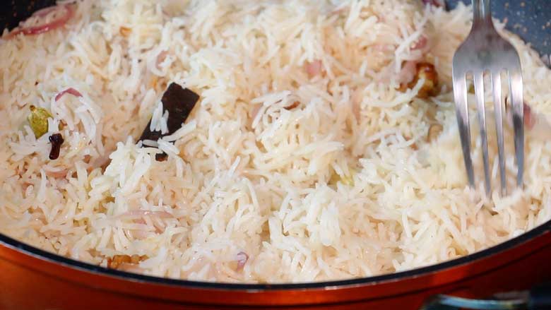 A metallic fork is seen fluffing the cooked Kerala ghee rice in an orange non stick pan. Seen also are whole spices in the rice - Kerala Ghee Rice with California Walnuts Recipe